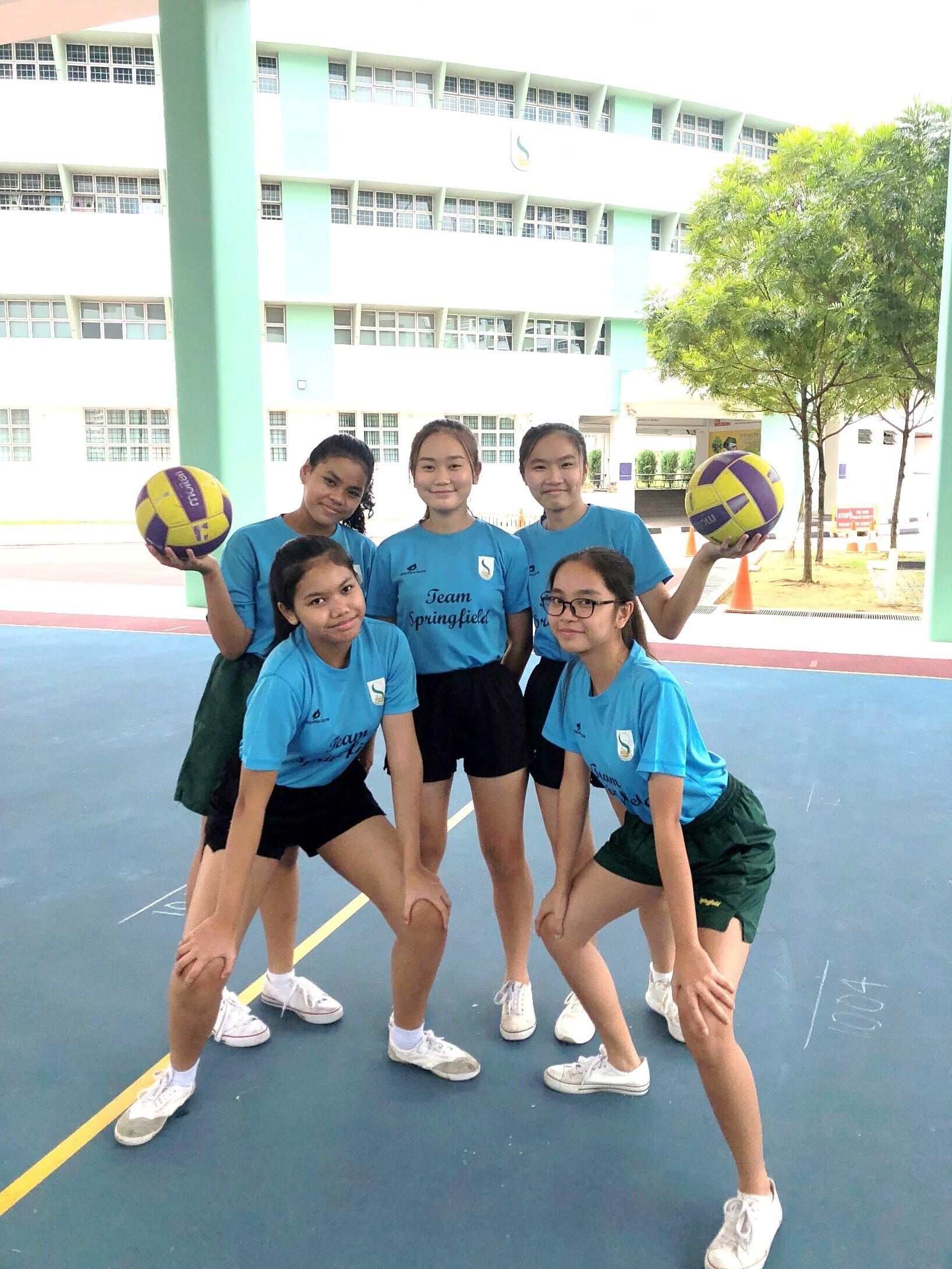 Five girls in blue Team Springfield shirts pose on a court, two holding yellow and purple volleyballs.
